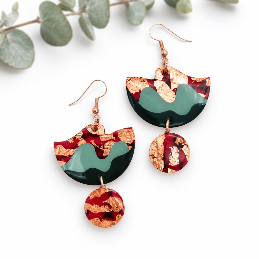 Abstract resin drop earrings with copper foil flakes, a dark green base, and red swirls, shown on a white background with eucalyptus leaves.