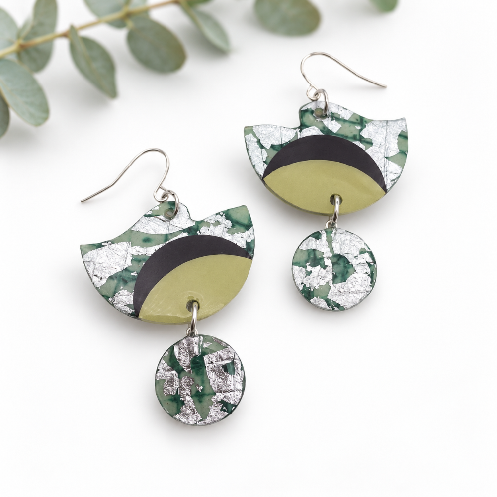 A pair of handmade dangle earrings with a green and silver marbled pattern, featuring a semi-circular top piece with a black curved accent, a dangling oval link, and a round drop charm, displayed on a white background with eucalyptus leaves.