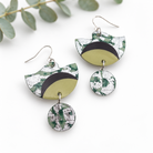 A pair of handmade dangle earrings with a green and silver marbled pattern, featuring a semi-circular top piece with a black curved accent, a dangling oval link, and a round drop charm, displayed on a white background with eucalyptus leaves.