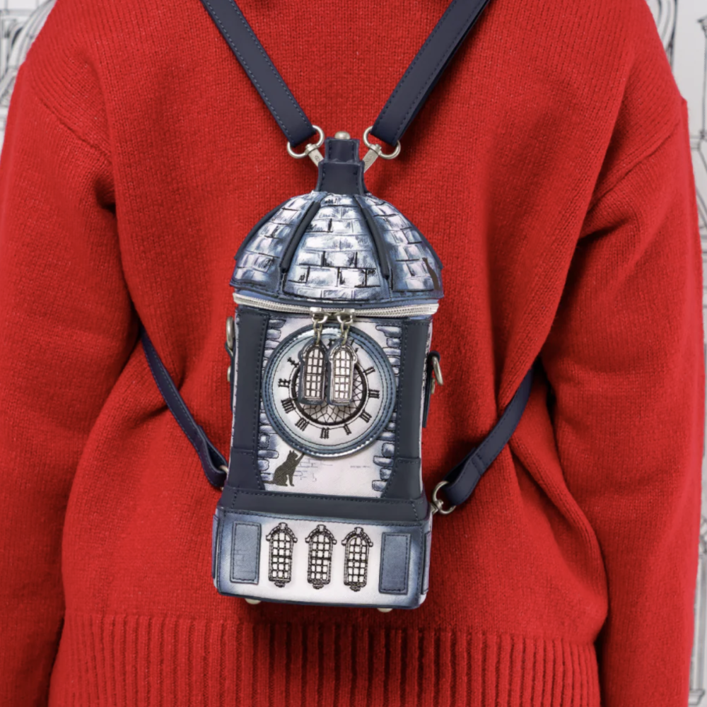 A person wearing a bright red knit sweater carries a small decorative backpack shaped like a clock tower, with dark navy straps and silver-toned detailing.