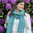 A woman wearing a teal blue knit scarf with white fringe, styled over a white button-down shirt and black pants, standing in front of a background of blooming purple flowers.