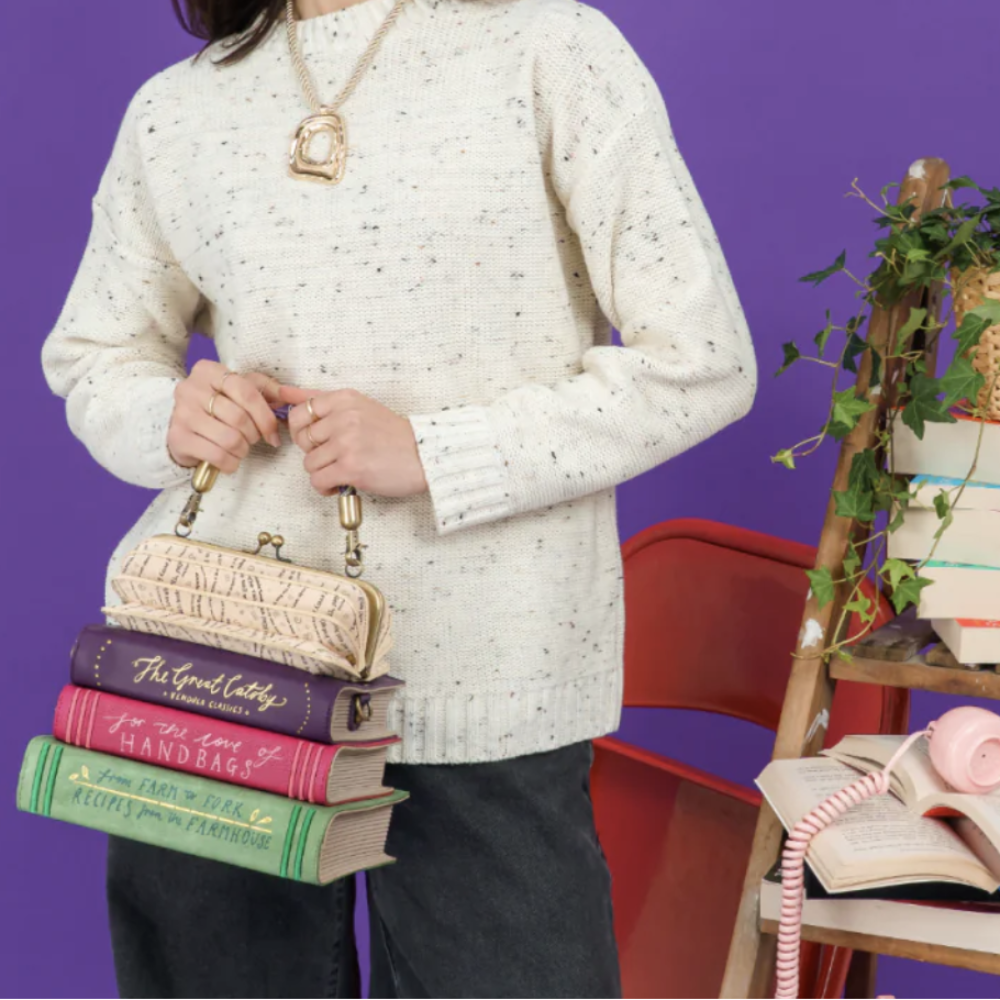 A person wearing a cream speckled knit sweater and dark pants holds a small clutch purse shaped like a stack of vintage books. The scene is styled with a purple wall, a red chair, and a wooden shelf holding books, a pink telephone, and a potted plant.
