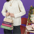 A person wearing a cream speckled knit sweater and dark pants holds a small clutch purse shaped like a stack of vintage books. The scene is styled with a purple wall, a red chair, and a wooden shelf holding books, a pink telephone, and a potted plant.