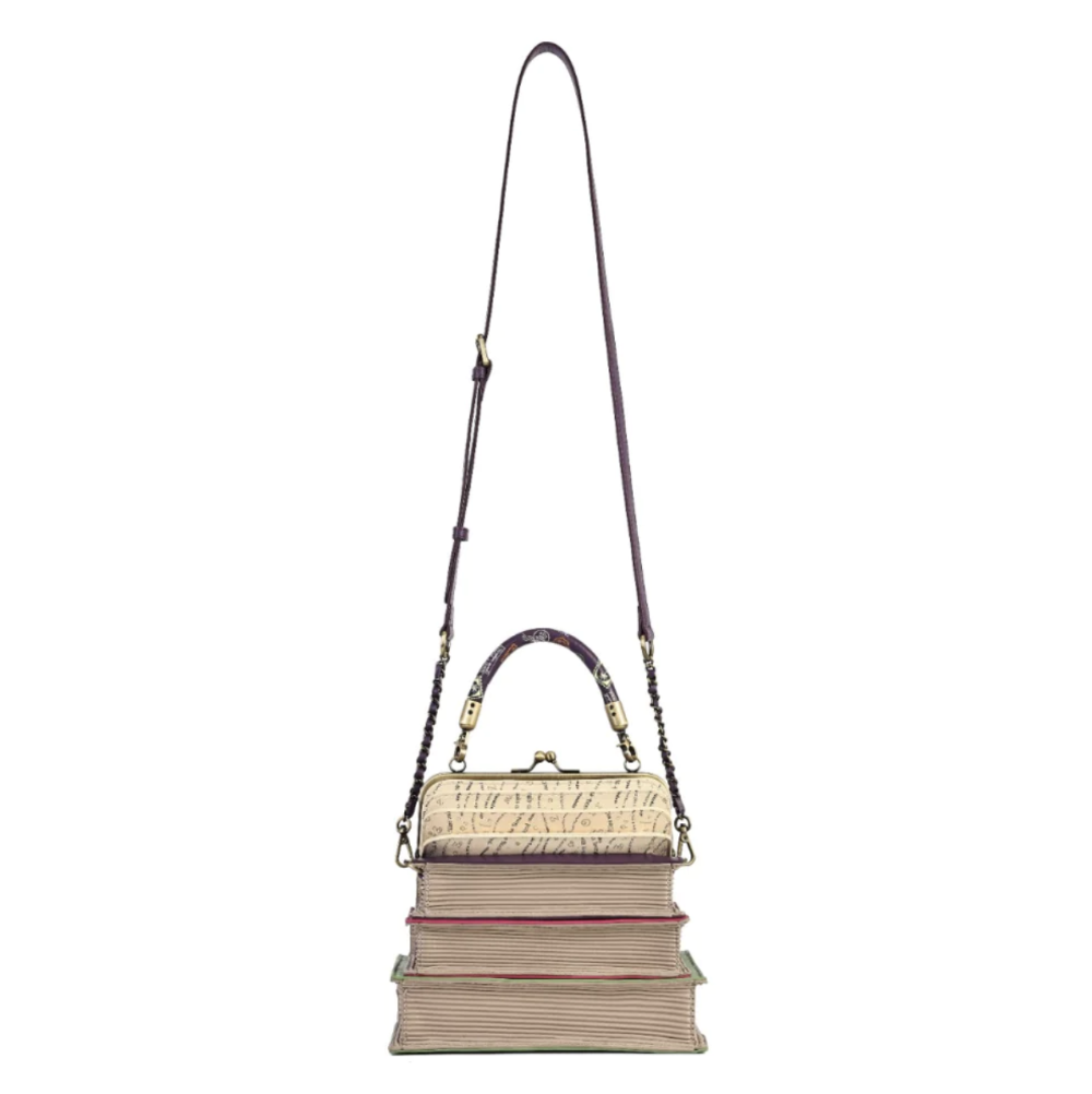 A handbag designed to look like a stack of vintage books, featuring a textured cream and gold faux-croc pattern on the top book, a beige and pink striped middle book, and a pink striped bottom book, with a dark purple shoulder strap and gold-tone hardware, shown on a plain white background.