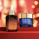 Two Estée Lauder skincare products displayed on a red surface against a warm blurred holiday background: the Advanced Night Repair serum bottle on the left and the Revitalizing Supreme+ cream jar on the right.