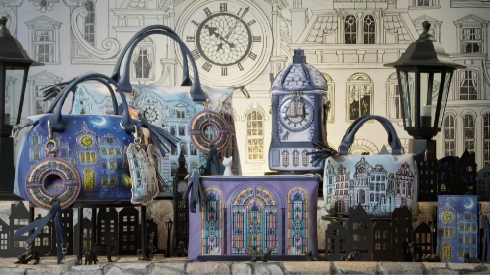 A collection of handbags and small decorative accessories featuring detailed illustrations of gothic houses, clocks, and windows in a whimsical village scene, displayed against a matching illustrated building backdrop.