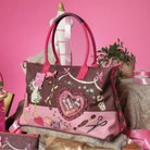 A pink and mauve patchwork tote bag with sewing and beauty-themed appliques, displayed on a styled pink set with a mannequin torso, ribbon, and small plant.