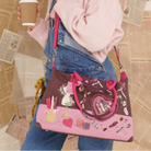 A person wearing a pink sweater and denim overalls carries a pink and brown decorative shoulder bag with cartoon-style charms and a heart-shaped pouch, while holding a coffee cup against a background of scattered newspaper pages.