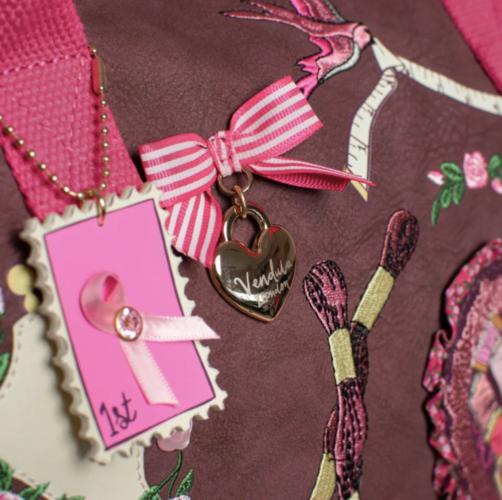 A close-up of a pink and white striped ribbon bow with a gold heart-shaped charm engraved with the brand name 'Vendula Paris'. The charm is attached to a pink ribbon and displayed on a dark brown patterned fabric alongside a pink commemorative card.